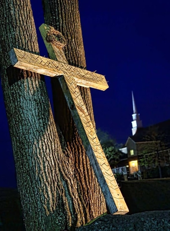 Illuminated wooden cross leaning against a tree at night with a church steeple in the background
